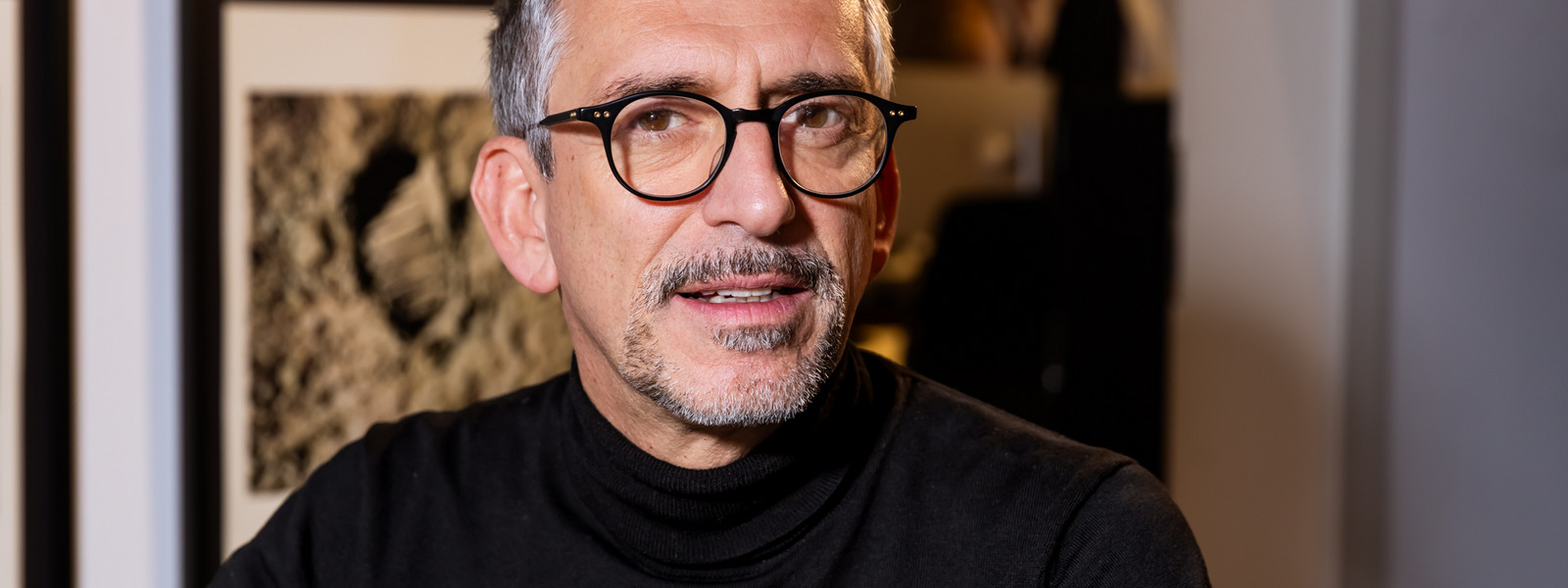 Joram Scher, Partner von Bachmann & Scher, mit Brille und schwarzem Rollkragenpullover vor gerahmten Fotografien in den Münchner Geschäftsräumen