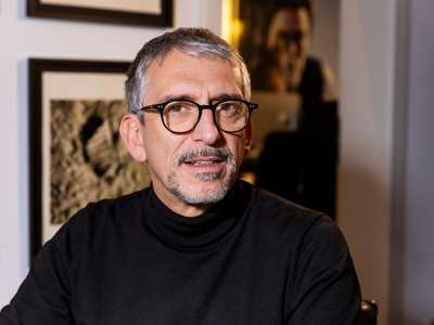 Joram Scher, Partner von Bachmann & Scher, mit Brille und schwarzem Rollkragenpullover vor gerahmten Fotografien in den Münchner Geschäftsräumen