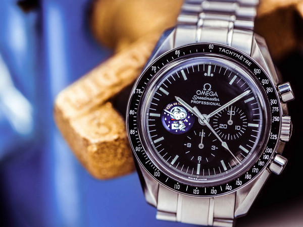 Omega Speedmaster Professional mit schwarzem Zifferblatt, schwarzer Tachymeter-Lünette und Snoopy-Hilfszifferblatt am Stahlarmband vor unscharfem Goldbarren